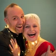 251216_Lametta.jpg Ein fröhliches Paar mittleren Alters lächelt, umgeben von einer Lichterkette vor grünem Hintergrund.