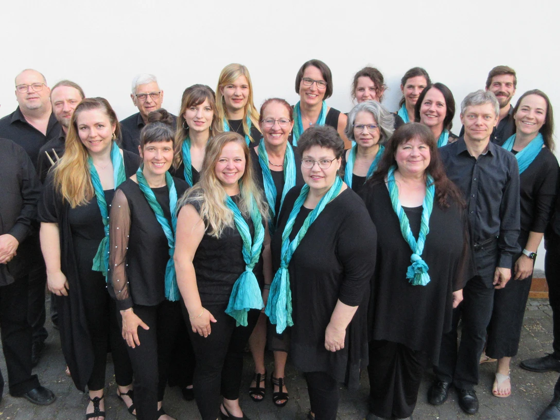 251224_StGospel.jpg Eine Gruppe von 19 Sängern in schwarz und türkis schalbekleidet vor einer hellen Wand.A group of 19 singers dressed in black and turquoise shawls in front of a light-colored wall.Skupina 19 zpěvaček oblečených do černých a tyrkysových šátků před světlou stěnou.Grupa 19 śpiewaków ubranych w czarne i turkusowe szale przed jasną ścianą.Een groep van 19 zangers gekleed in zwarte en turquoise sjaals voor een lichtgekleurde muur.Un gruppo di 19 cantanti vestiti con scialli neri e turchesi davanti a una parete chiara.