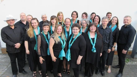 A group of 19 singers dressed in black and turquoise shawls in front of a light-colored wall.