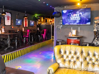 Lost Level entrance area Im Innenbereich eines Gaming-Cafés reflektieren bunte Lichter auf einem Boden aus blauen Fliesen. Eine Reihe von Tischen steht entlang der Seite, flankiert von schwarzen und pinken Stühlen unter mehreren Monitoren.Inside a gaming café, colorful lights reflect on a floor of blue tiles. A row of tables stands along the side, flanked by black and pink chairs under several monitors.