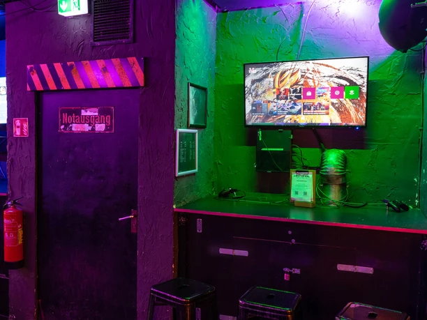 Gaming Area PS4 & XBOX A gaming zone with PS4 and Xbox consoles, gloomily lit, with bar stools in front of display screens.