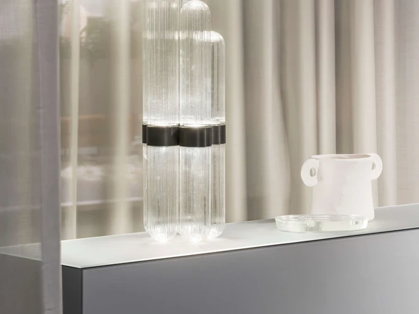 idd cologne: Koelnmesse GmbH / Constantin Meyer Ein modernes Interieur mit schlichtem, grauem Regal, darauf eine elegante, glänzende Glaslampe.