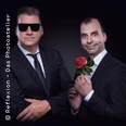 Zwei Männer in Anzug posieren vor dunklem Hintergrund, einer hält eine rote Rose, der andere trägt Sonnenbrille.