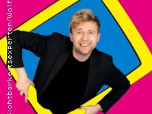 Ein Mann mit blondem Haar und schwarzem Jackett lächelt und schaut durch einen gelben, schrägen Rahmen.