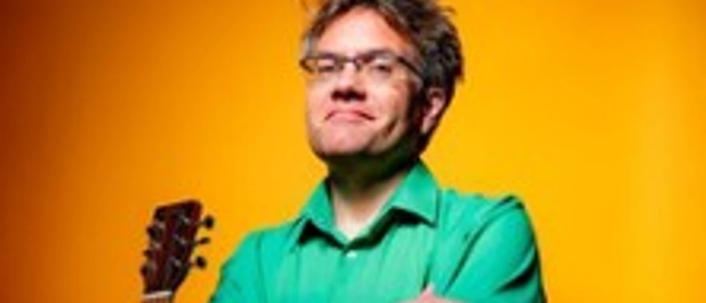 Ein Mittelalter Mann mit Brille im grünen Hemd posiert verschmitzt mit Gitarre und orangefarbenem Hintergrund.