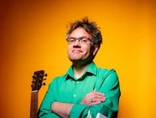 Ein Mittelalter Mann mit Brille im grünen Hemd posiert verschmitzt mit Gitarre und orangefarbenem Hintergrund.