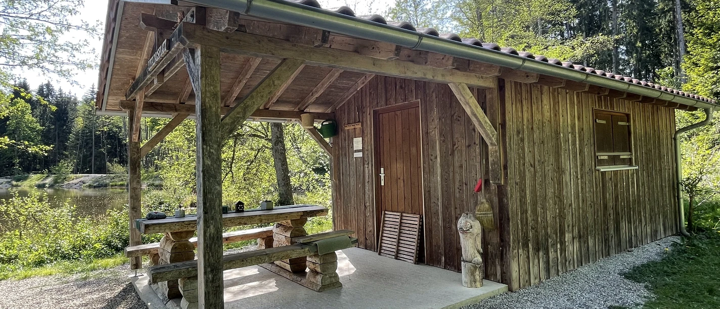 rustikales Holzgebäude mit überdachtem Sitzbereich, umgeben von Wald und einem stillen Weiher