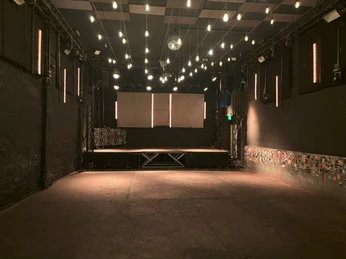Helio37 Mainfloor Leerer Veranstaltungsraum mit Bühne, umgeben von dunklen Wänden und Lichterketten an der Decke.Empty event room with stage, surrounded by dark walls and fairy lights on the ceiling.