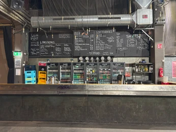 Live Music Hall Eine Bar mit Kreidetafel-Menü, Kühlschränken voller Getränke und einem industriellen Deckendesign.A bar with a chalkboard menu, fridges full of drinks and an industrial ceiling design.