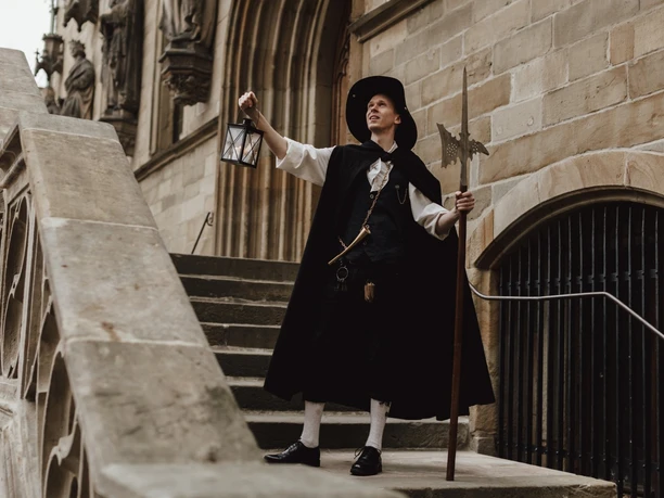 Nachtwächter (2).jpg Eine Nachtwächterin führt mit Laterne und Hellebarde geschichtsbewusste Besucher über eine Steintreppe.