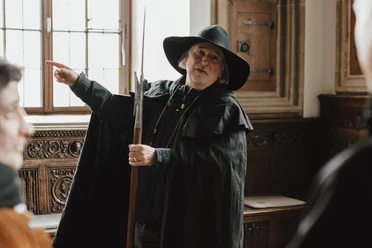 Nachtwächter (Hauptbild).jpg Een nachtwaker in historisch kostuum geeft uitleg aan bezoekers in een traditioneel ingerichte kamer.