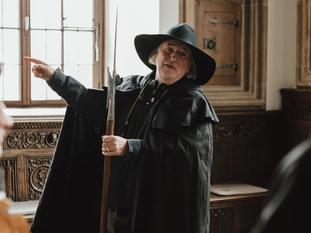 Nachtwächter (Hauptbild).jpg Ein Nachtwächter im historischen Kostüm erklärt Besuchern in einem traditionell eingerichteten Raum Details.