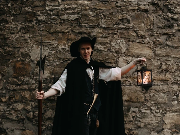 Beschwipster Nachtwächter (2).jpg A night watchman in traditional dress leans against an old stone wall and holds a lantern.