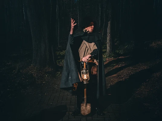 Galgen, Gräber & Ganoven (3).jpg Ein Mann in historischer Kleidung mit Umhang und Laterne steht in einem dunklen Wald auf einem Weg.