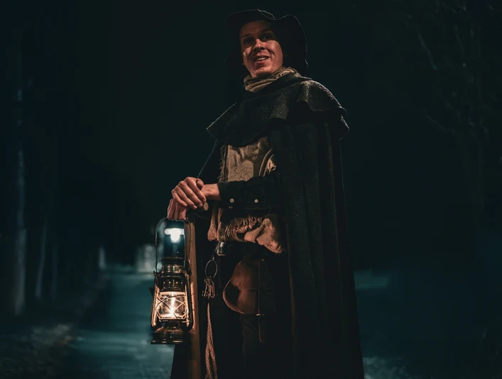 Galgen, Gräber & Ganoven (Hauptbild).jpg Ein Nachtwächter in traditionellem Gewand hält eine Laterne und steht auf einem dunklen Weg.A night watchman in traditional garb holds a lantern and stands on a dark path.En nattevagt i traditionel klædedragt holder en lanterne og står på en mørk sti.Een nachtwaker in traditioneel gewaad houdt een lantaarn vast en staat op een donker pad.