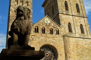 Altstadt-Highlights (Hauptbild).jpg Steinerne Skulptur eines Löwen vor einer historischen Kirche mit zwei Türmen und einer großen Uhr.