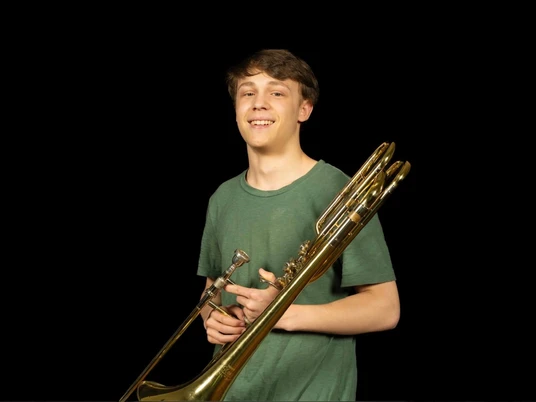 Solist-Carl-Rabbels-Schlossorchester-Oldenburg.jpg Ein junger Musiker mit grünem Shirt hält eine Posaune und lächelt vor dunklem Hintergrund.