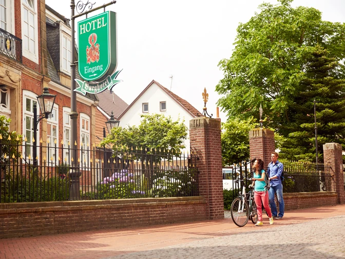 Zwei Radfahrer gehen vor dem historischen Burghotel Haselünne entlang einer Baumallee im Emsland.