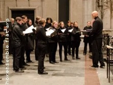 Chorensemble singt in einer Kirche mit gotischen Bögen im Hintergrund, dirigiert von einer Person.