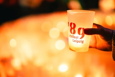Hand holding an illuminated cup with the inscription "Lichtfest Leipzig '89" in front of a blurred candlelight background.