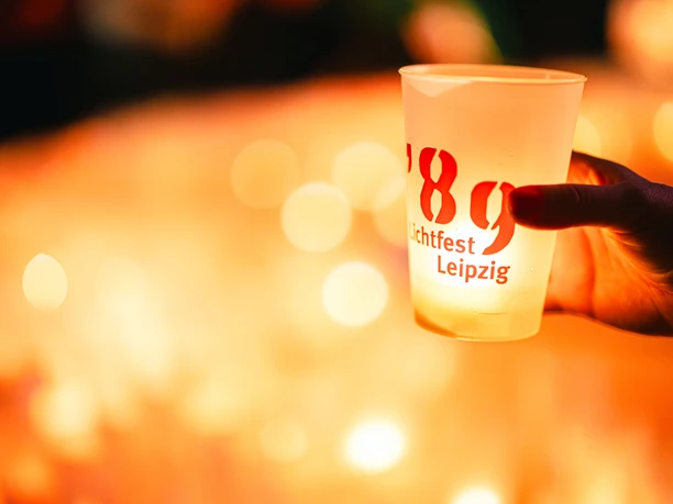 Hand hält beleuchteten Becher mit Aufschrift "Lichtfest Leipzig '89" vor unscharfem Kerzenlicht-Hintergrund.