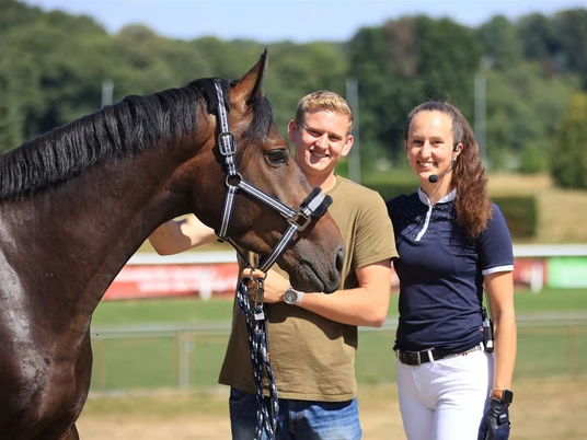 Zwei Menschen stehen lächelnd neben einem Pferd auf einem Rasenplatz an einem sonnigen Tag.