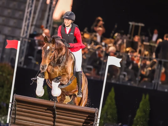 Reiterin springt mit einem braunen Pferd über ein Hindernis, im Hintergrund ein Orchester.