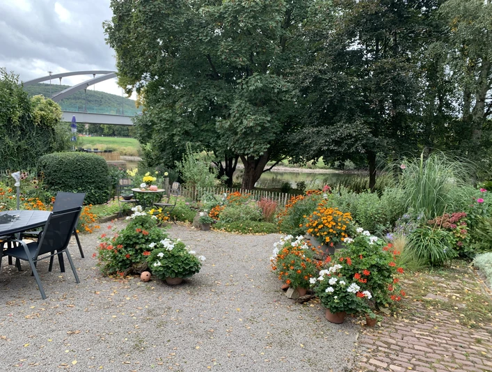 Garten und Weserblick der Ferienwohnung Rad und Ruhe Idyllischer Garten mit Blumen und Baumbestand, Gartenmöbeln, im Hintergrund Blick auf die Weser und Weserbrücke