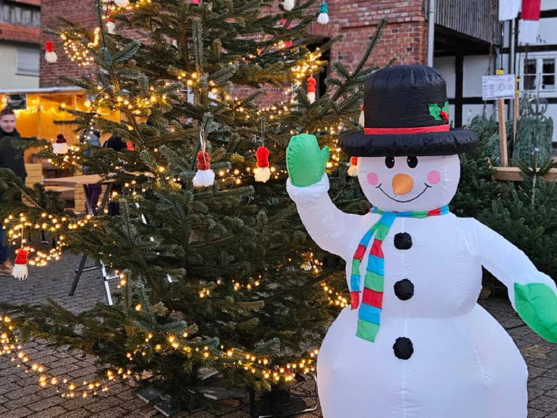 schneemann.jpg Aufblasbarer Schneemann mit Hut und Schal neben festlich beleuchtetem Weihnachtsbaum auf Markt.