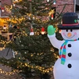 schneemann.jpg Aufblasbarer Schneemann mit Hut und Schal neben festlich beleuchtetem Weihnachtsbaum auf Markt.