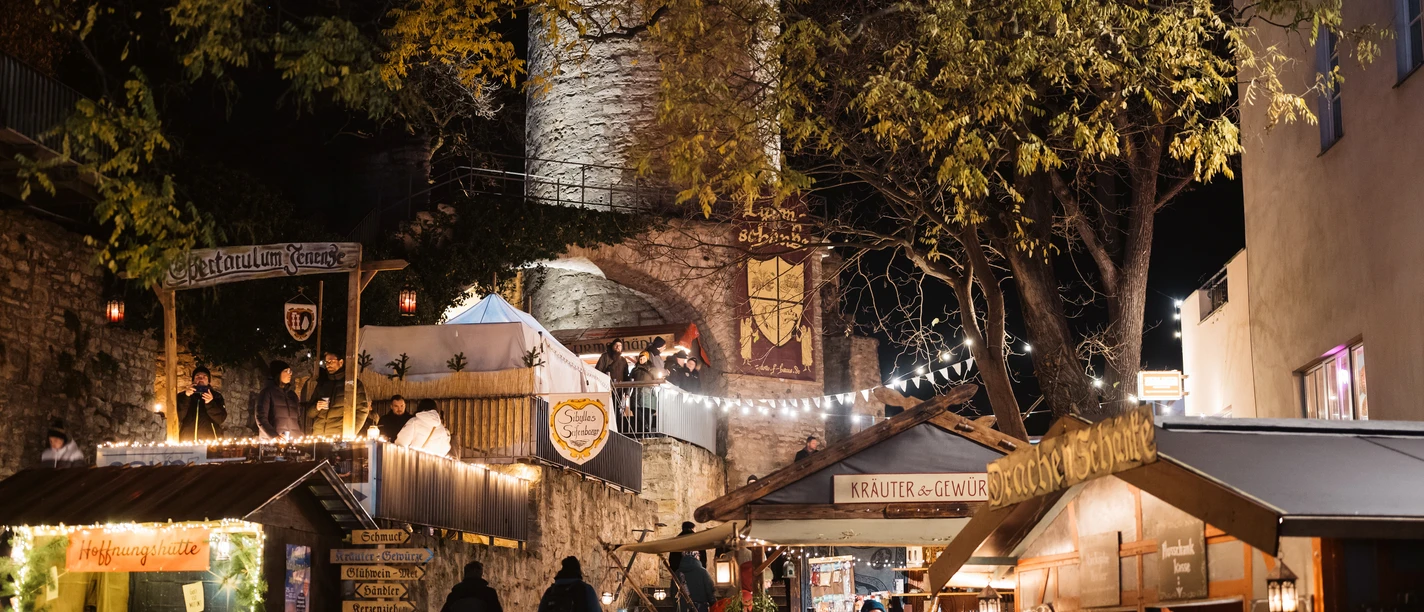 Aufnahme vom Historischen Weihnachtsmarkt mit Ständen, Feuerstellen sowie vielen Besucher:innen und einem historischem Wehturm im Hintergrund.