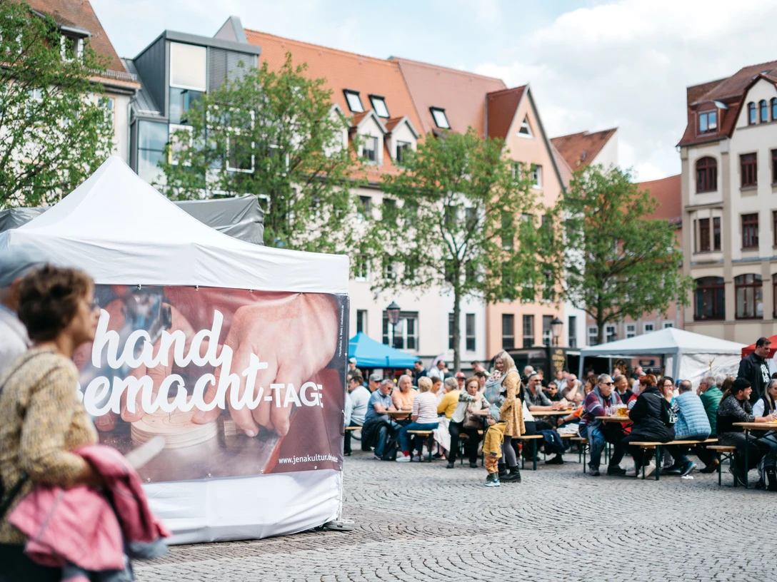HandgemachtTage©JenaKultur_CWorsch+(106).jpg Zwei Menschen laufen an einem Pavillon vorbei, an dem ein Banner gehängt wurde, an dem darauf steht "handgemacht-Tage", im Hintergrund sitzen mehrere Menschen auf Bierbänken.