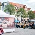 HandgemachtTage©JenaKultur_CWorsch+(106).jpg Zwei Menschen laufen an einem Pavillon vorbei, an dem ein Banner gehängt wurde, an dem darauf steht "handgemacht-Tage", im Hintergrund sitzen mehrere Menschen auf Bierbänken.