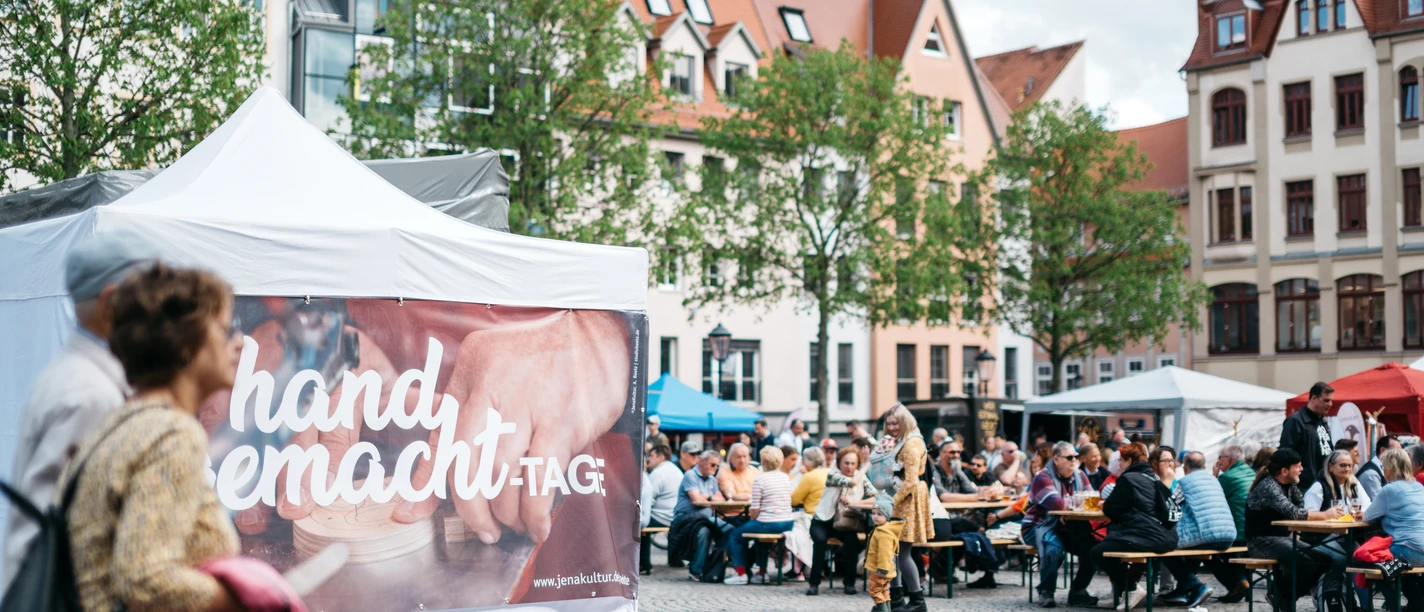 HandgemachtTage©JenaKultur_CWorsch+(106).jpg Zwei Menschen laufen an einem Pavillon vorbei, an dem ein Banner gehängt wurde, an dem darauf steht "handgemacht-Tage", im Hintergrund sitzen mehrere Menschen auf Bierbänken.