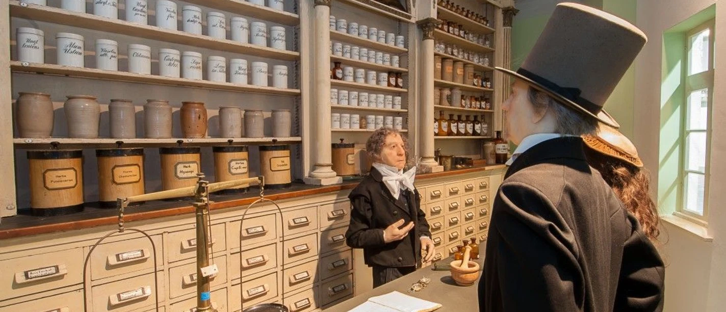 Historische Apotheke mit traditionellen Apothekerschränken und Gefäßen. Eine Wachsfigur steht im historischen Kostüm vor der Theke und eine dahinter.