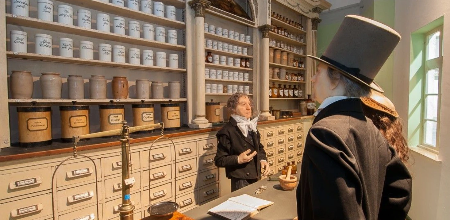 Historische Apotheke mit traditionellen Apothekerschränken und Gefäßen. Eine Wachsfigur steht im historischen Kostüm vor der Theke und eine dahinter.