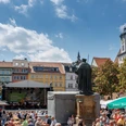 JenaerFrühlingsmarkt©JenaKultur_Moebius20249643c.jpg Szenerie des Jenaer Frühlingsmarktes mit Bühne, vielen Menschen, die sich um die Statue des Hanfriedes versammeln.
