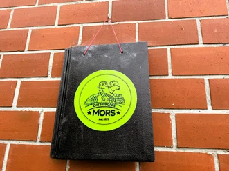 Dat Hofcafé Schwarzes Schild mit rundem, gelbgrünem Aufkleber, darauf Zeichnung von Bäumen und Haus, Text 'DAT HOFCAFÉ MORS Seit 2021', an roter Schnur an roter Backsteinwand hängend.