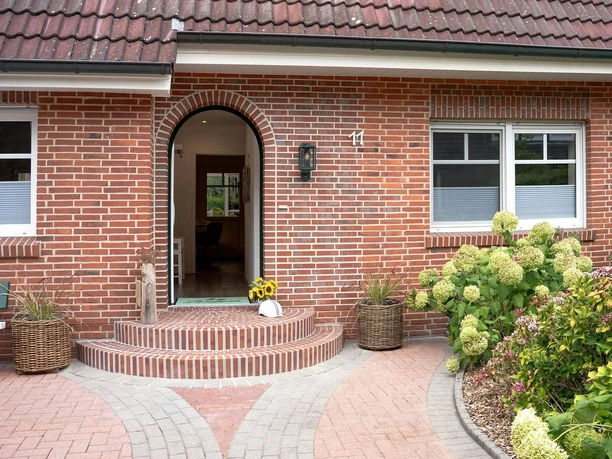landhuus-geeste-13.jpg Eingangsbereich eines roten Klinkerhauses mit Rundbogentür, Blumen und gepflegtem Gartenweg.