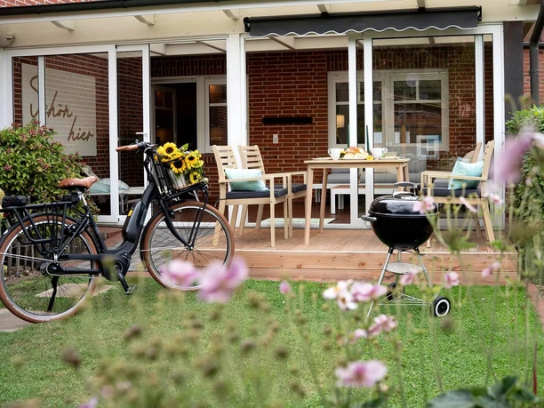 landhuus-geeste-24.jpg Terrasse mit Holztisch, Gartenstühlen und Grill vor rotem Klinkerhaus, daneben Fahrrad mit Sonnenblumenkorb.