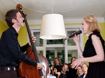Musik in den Häusern der Stadt Ein intimes Konzert in einem gemütlichen Wohnzimmer: Ein Kontrabassist und eine Sängerin treten vor einem entspannten Publikum auf, das in Sesseln und auf einem Sofa sitzt. Der Raum bietet eine freundliche Atmosphäre mit gelben Wänden und einem großen Fenster im Hintergrund.An intimate concert in a cozy living room: a double bass player and a singer perform in front of a relaxed audience sitting in armchairs and on a sofa. The room has a friendly atmosphere with yellow walls and a large window in the background.
