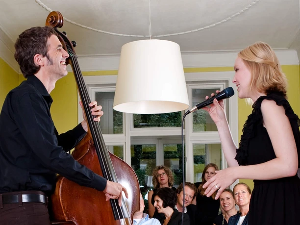 Musik in den Häusern der Stadt An intimate concert in a cozy living room: a double bass player and a singer perform in front of a relaxed audience sitting in armchairs and on a sofa. The room has a friendly atmosphere with yellow walls and a large window in the background.