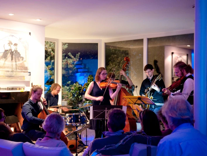 Musik in den Häusern der Stadt 2017 Eine Gruppe von Musiker*innen spielt konzentriert in einem modern beleuchteten Raum vor einem sitzenden Publikum.A group of musicians plays in a modern, well-lit room in front of a seated audience.