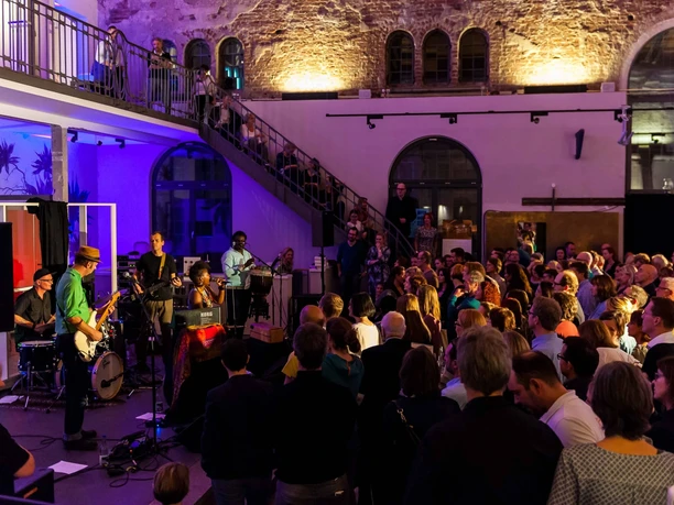 Musik in den Häusern der Stadt Ein Ensemble von Musiker*innen spielt vor einem faszinierten Publikum in einer stimmungsvoll beleuchteten Halle.
