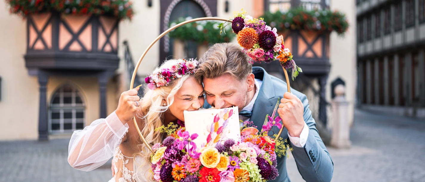 Hochzeitmesse Brautpaar mit Blumenkranz und Hochzeitstorte vor Fachwerkhäusern in Wernigerode.