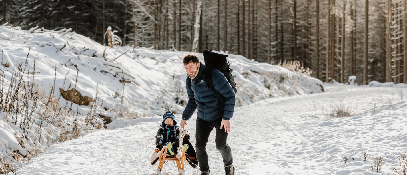 Ein Mann zieht ein Kind auf einem Holzschlitten durch den verschneiten Wald bei Schierke.