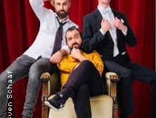 Drei Männer posieren vor einem roten Vorhang; zwei stehen, einer sitzt lässig vorne.
