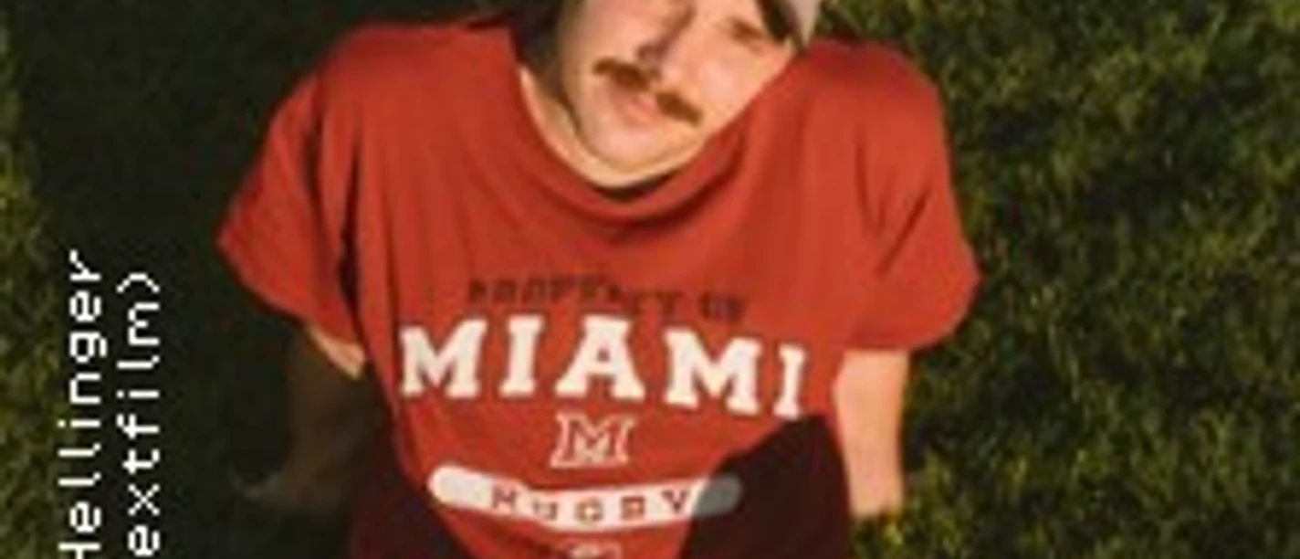 Ein junger Mann mit Schnurrbart und Baseballkappe sitzt entspannt auf grünem Rasen, Blick zur Kamera.