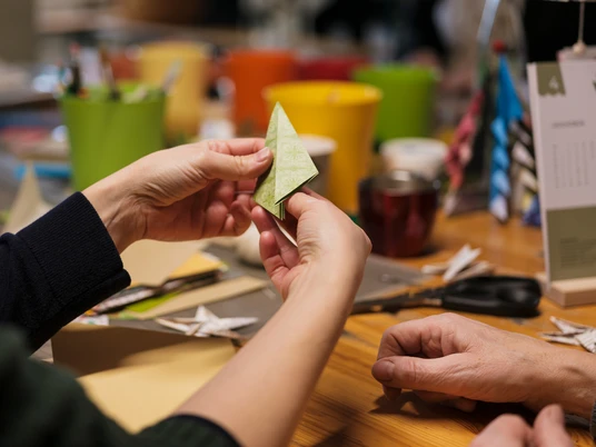 Roßweiner Weihnachtsmarkt - Weihnachten in der Region Leipzig Zwei Hände falten vorsichtig ein grünes Origamipapier zu einem Papierflugzeug, auf einem Tisch voller bunter Bastelutensilien