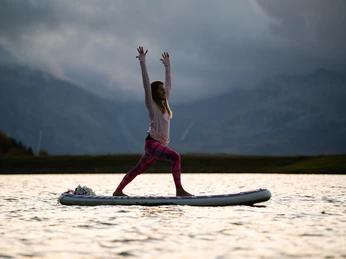 Yoga on SUP in the evening SUP Yoga Bettmersee im Sommer mit Frau in Kriegerhaltung bei stimmungsvoller Abendsonne und Bergsilhouette im HintergrundSUP Yoga Bettmersee in summer with woman in warrior pose with atmospheric evening sun and mountain silhouette in the backgroundSUP Yoga Bettmersee en été avec une femme en position de guerrière sous un soleil couchant évocateur et une silhouette de montagne en arrière-plan